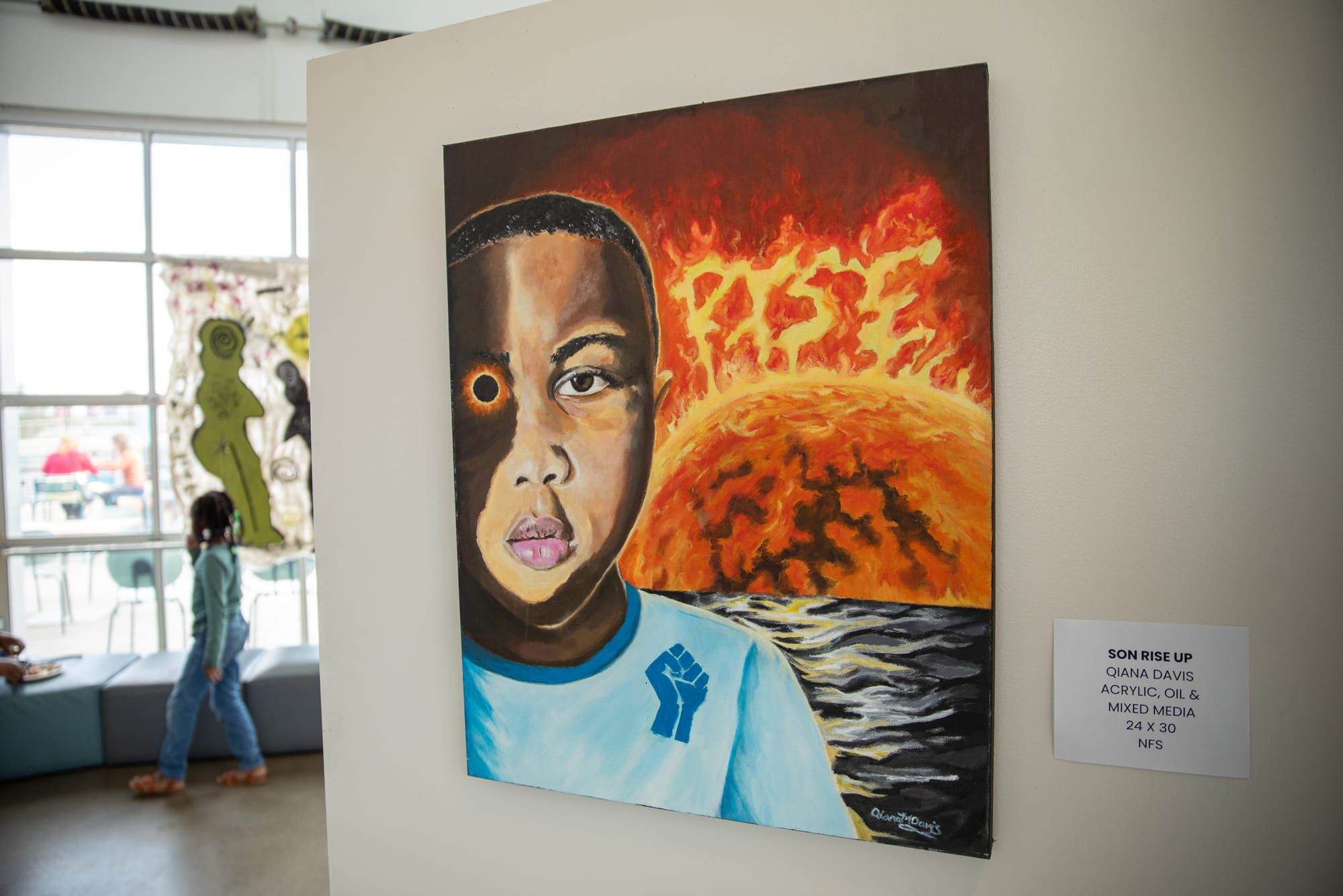 A painting of a young Black boy with a raised fist on his shirt, one eye glowing orange, stands before a fiery sunrise with the word "RISE" in the sky, displayed in a gallery.