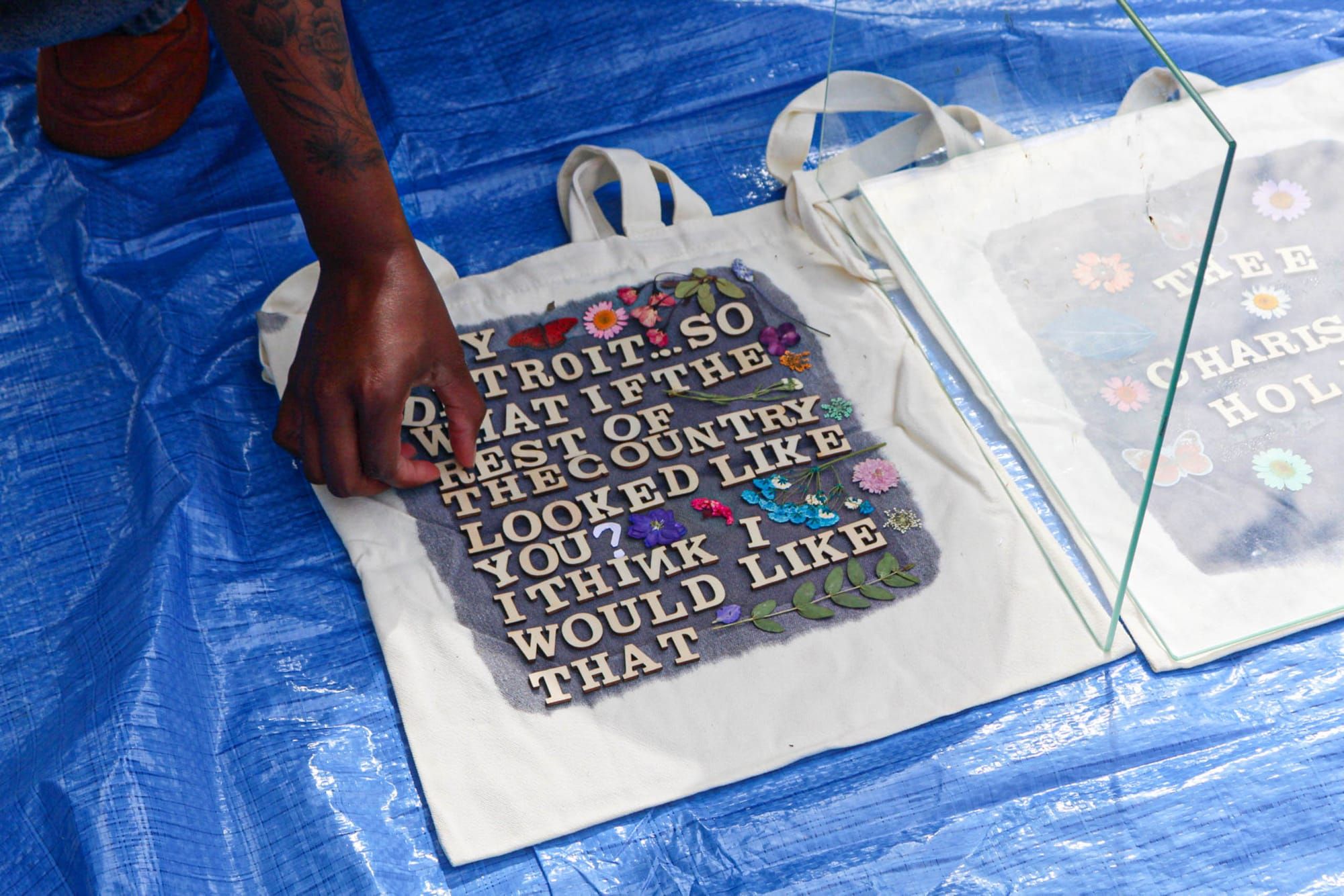 A hand arranges letters on a tote bag on a blue tarp that reads“Hey Detroit, what if the rest of the country looked like you? I think I would like that.”