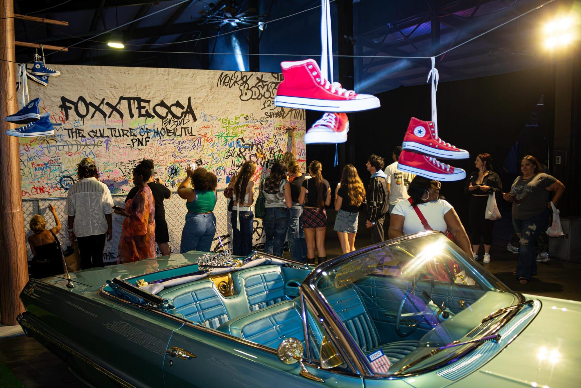 A crowd of people are facing a wall of graffiti behind a blue car, as shoes are tied to wires above.
