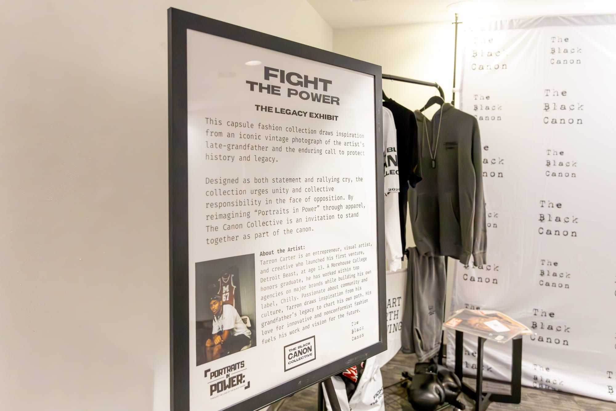 A white and black frame stands on an easel with the words “Fight the Power” a clothing rack and backdrop stands on the back. 