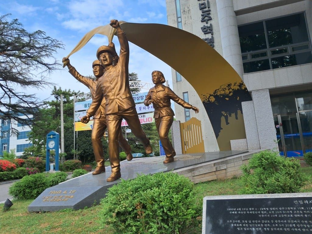 Larger than life-size bronze statue of three young adults running and shouting. They are carrying a long banner, which shows an illustration of a larger crowd.