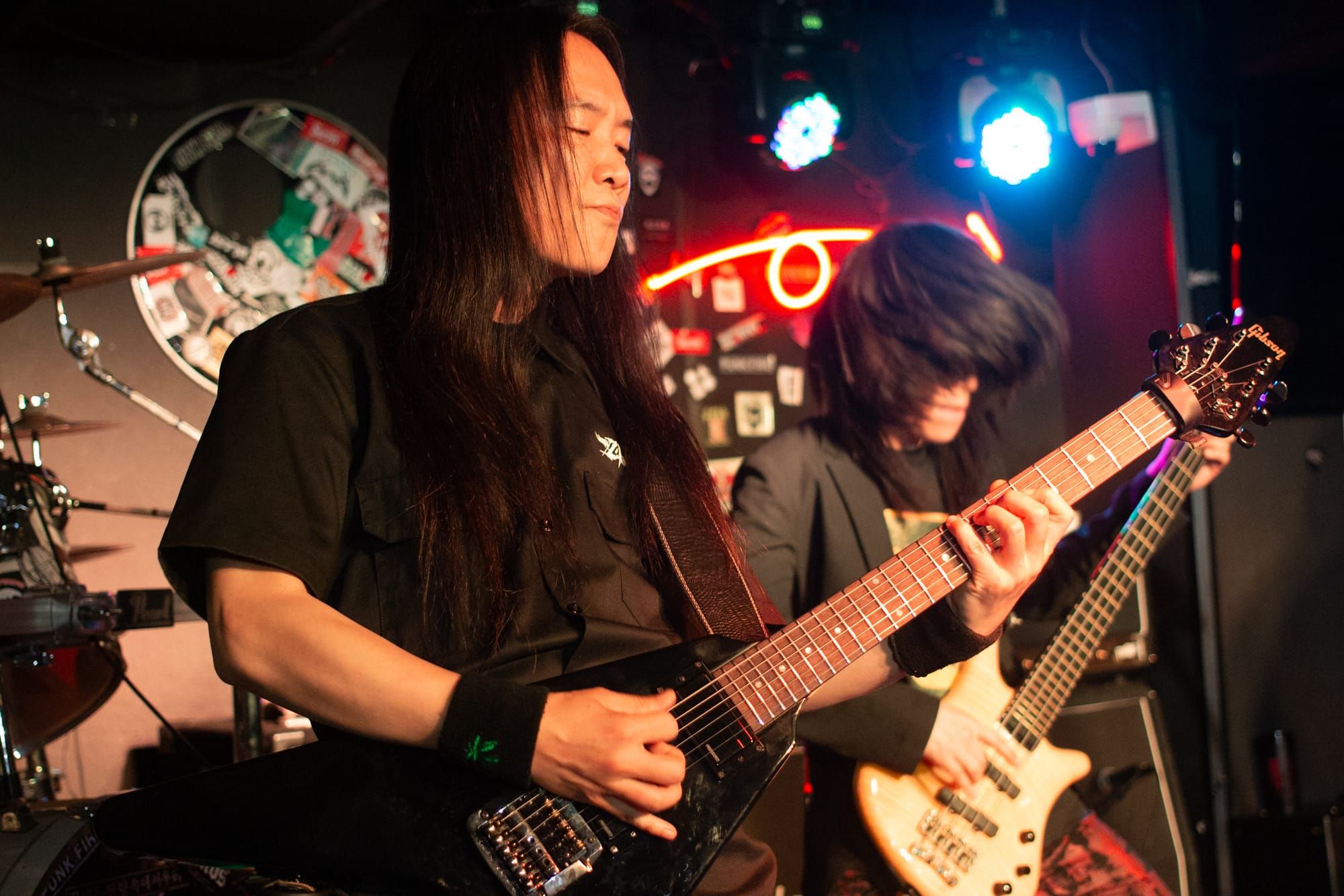 Lee Hee-Doo plays a guitar in a wide stance with a stoic expression. Behind him, bassist Han Seon-Jae flips his hair over his face while playing. Neon and stage lights illuminate them.