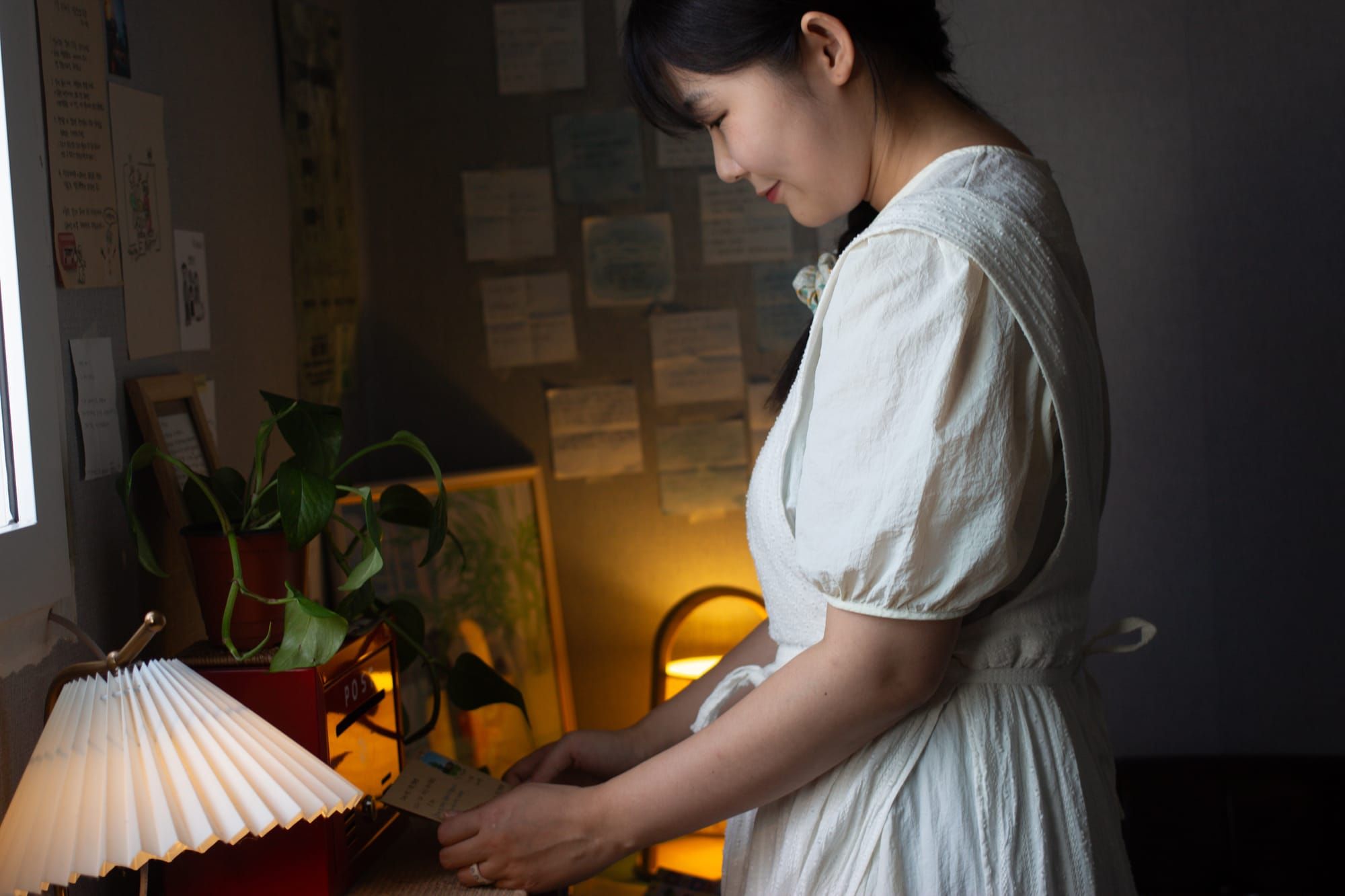 Seo Eun Ji, dressed in white, looks down at a desk with some lamps and plants.