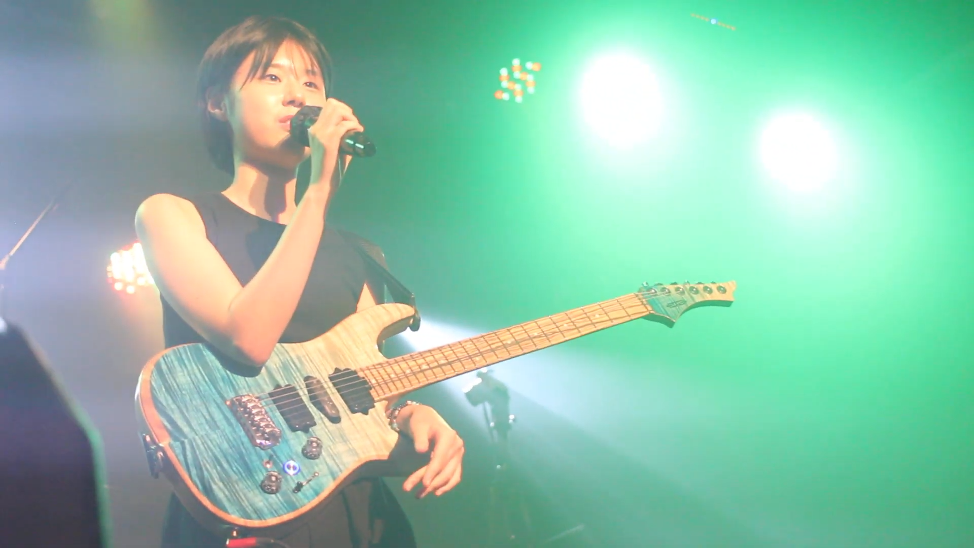 A girl with short hair holds a guitar with a beach design on it. She is speaking into the mic. Behind her are green stage lights.