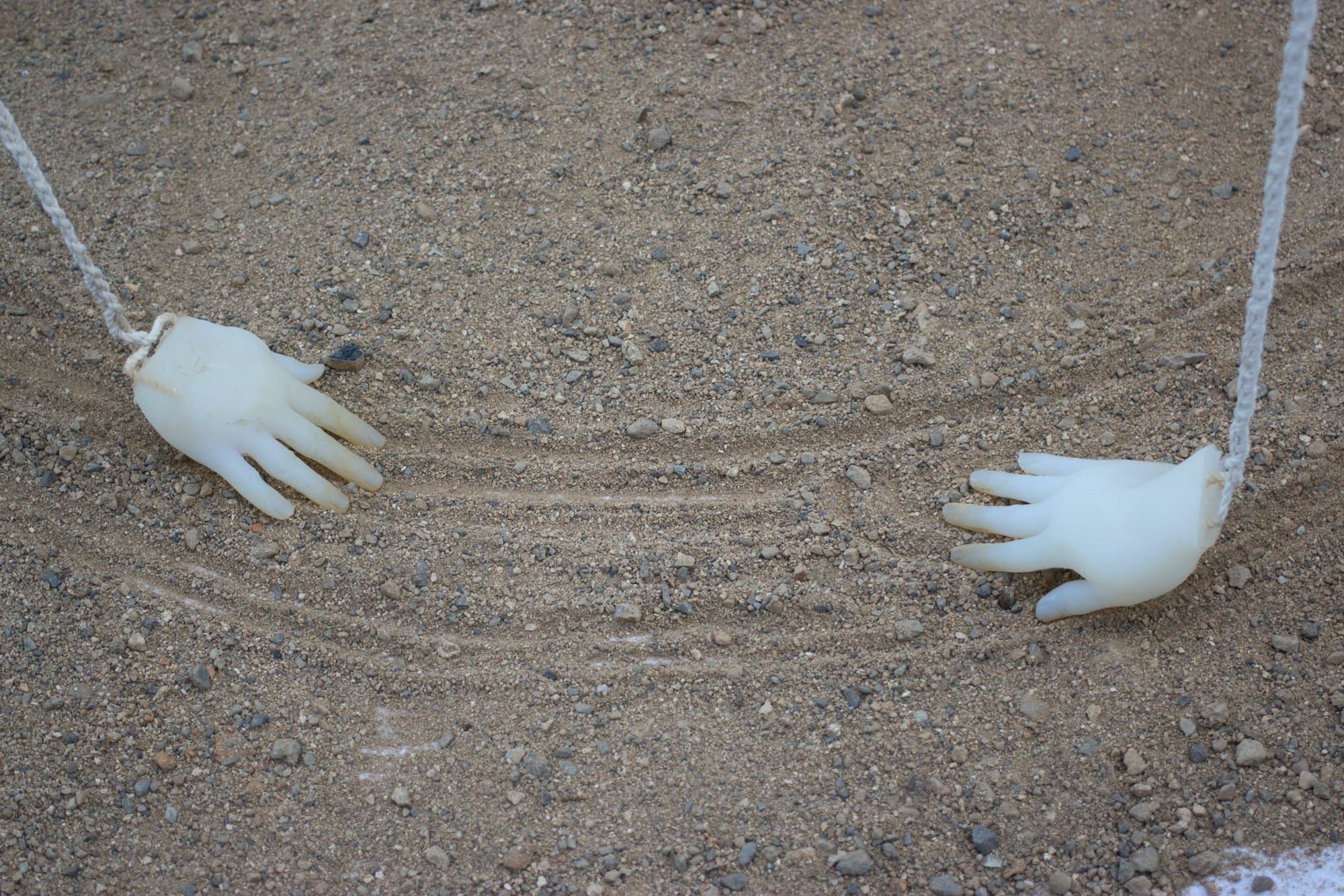Two translucent hand sculptures suspended by white ropes touch the sandy ground, leaving grooves in their paths. Their fingers are pointed toward each other.