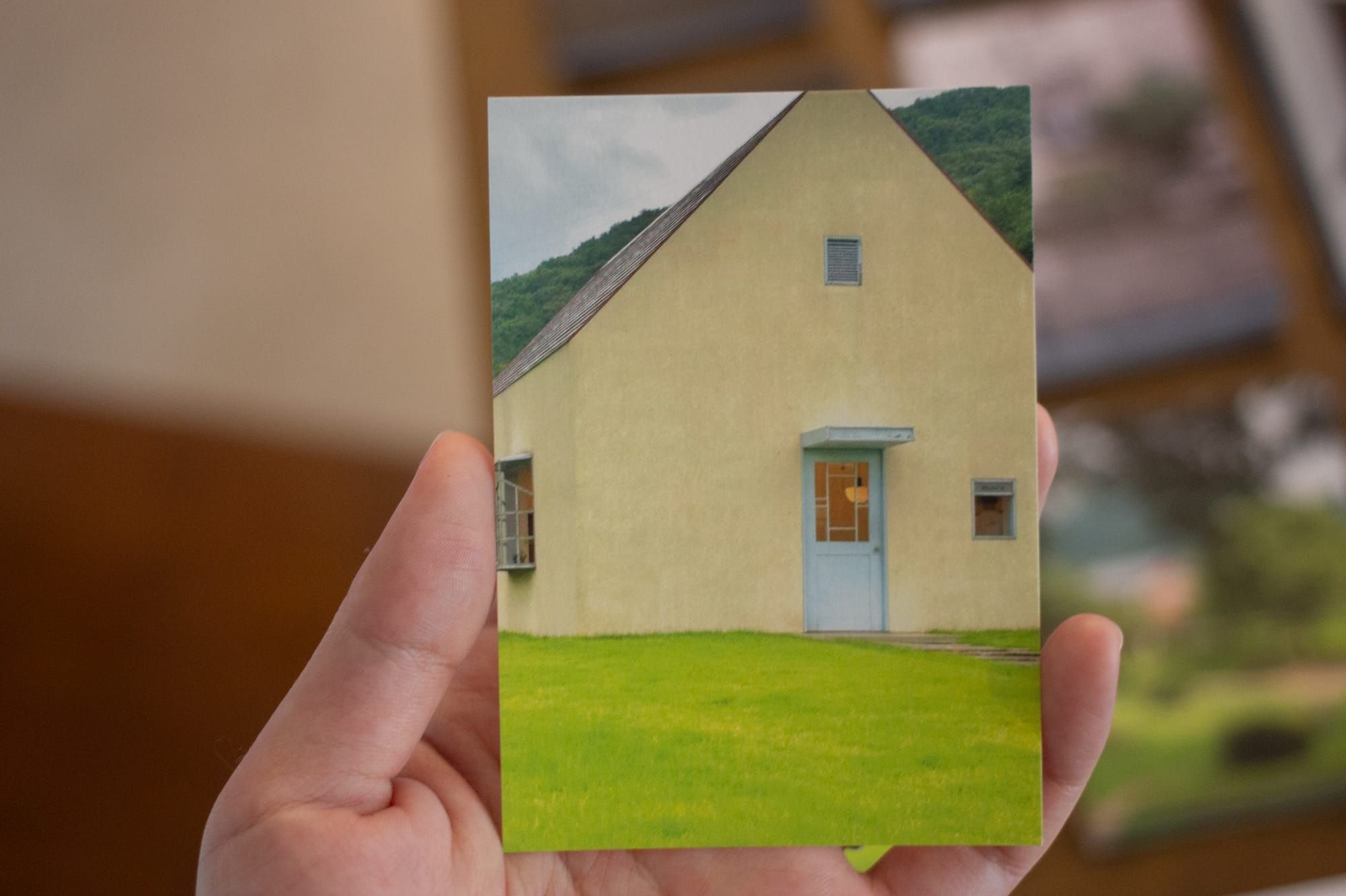 A hand holding a photograph of a simple triangular house in a bright green field. The house has small windows and a light blue door.