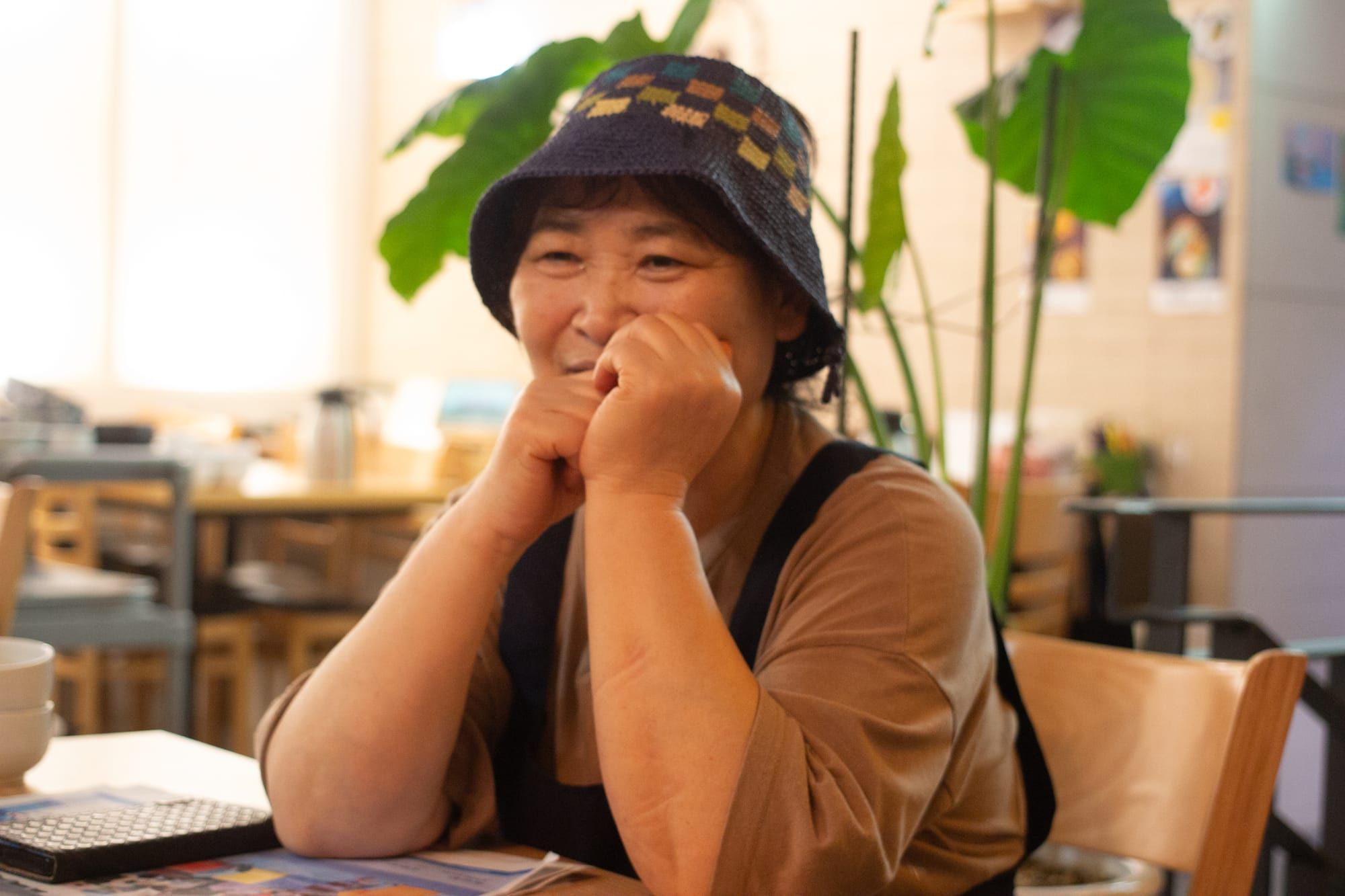 Kim Suha holds her hands by her face, thinking. She is sitting at a table in her restaurant, wearing a hat with different colored squares. Behind her is a large green plant.