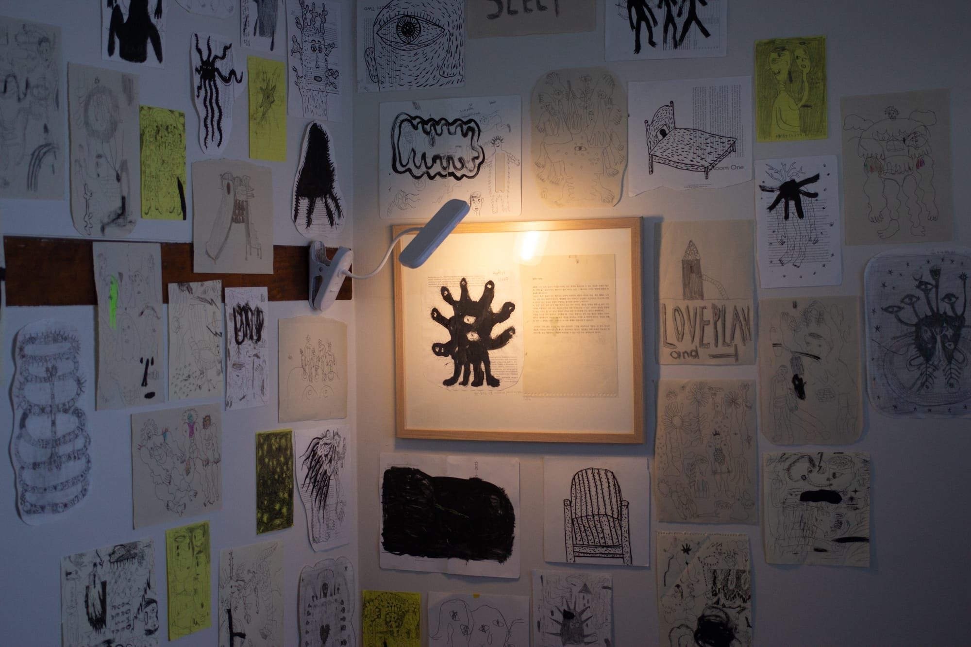 An interior corner filled with drawings on loose pieces of paper. A lamp illuminates a framed two-page spread featuring a monster with many eyeballs and a printed text passage.