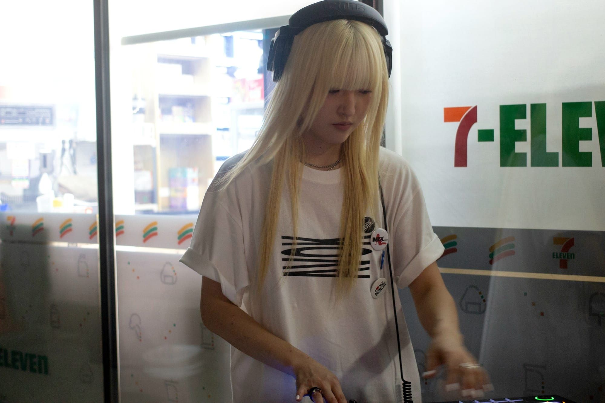 DJ Oto twists dials on a DJ deck with lit-up buttons. She has rings and bleached hair with straight-cut bangs. She is wearing big headphones.