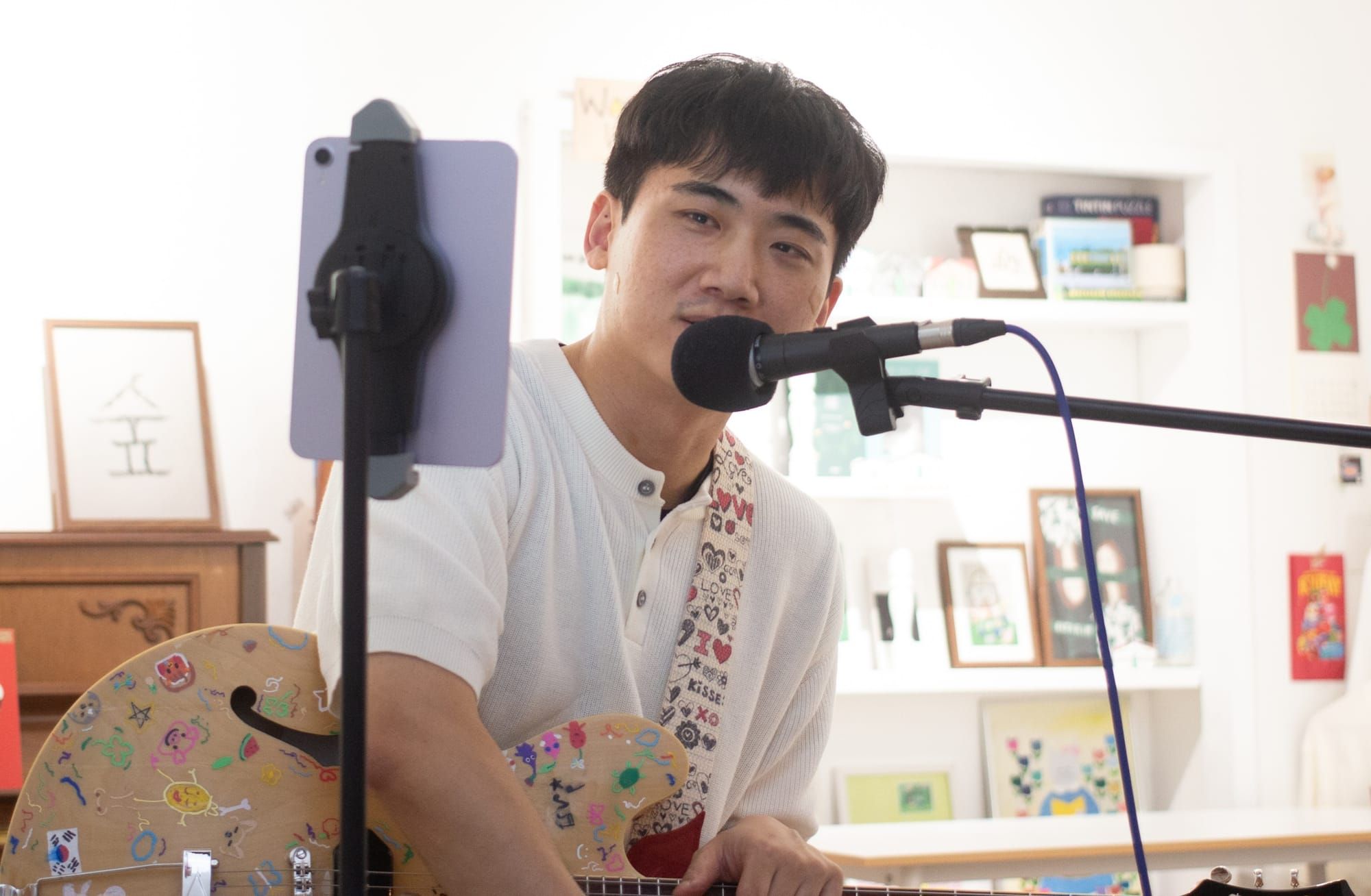 xx sits at a mic with a guitar covered in colorful drawings, with a guitar strap with prints of words like "Love," Kisses" and "XO." Behind are framed artworks.