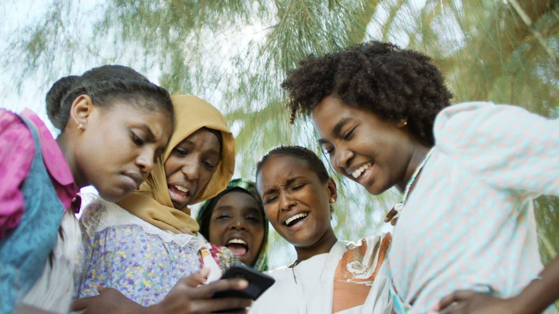 Five girls look at a phone screen in disgust and laughter. Behind is a tree; they are outside.