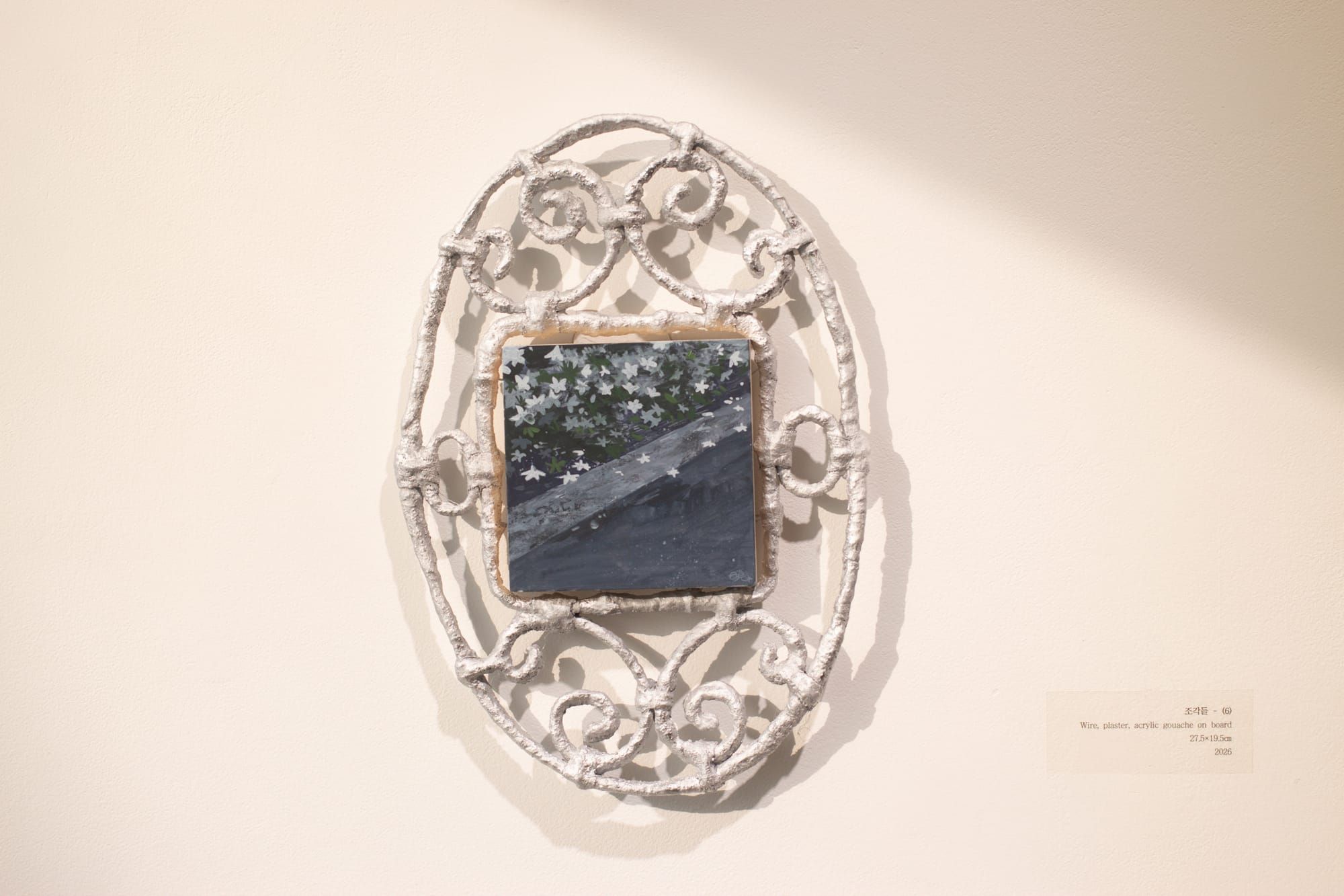 A small square painting framed by a textured white wire oval with curlicues. The painting depicts tiny white flowers and a cool gray solid surface, maybe asphalt.