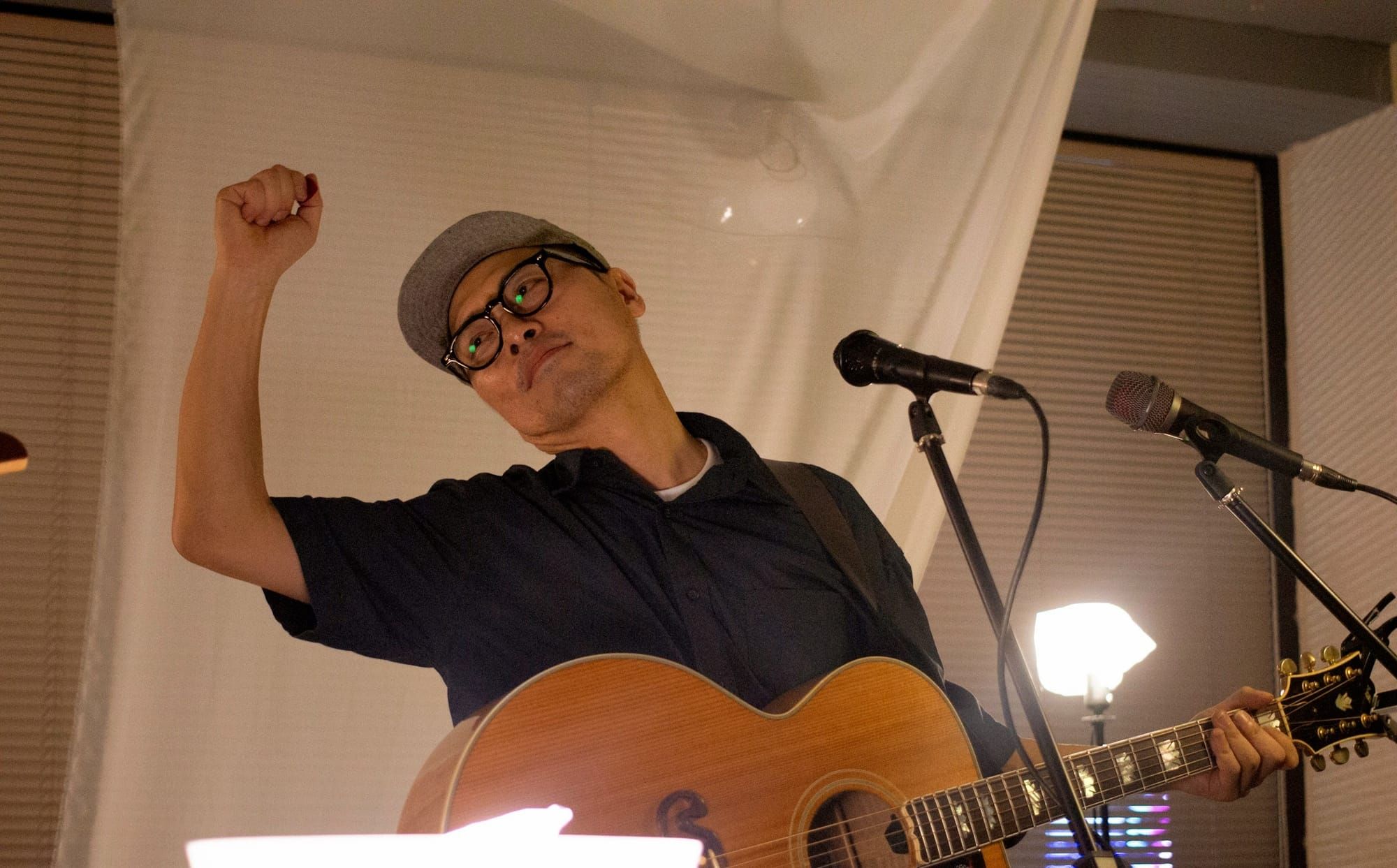 Lim Woong, standing with an acoustic guitar in front of two mics and lamps, in front of a thin white curtain, poses with one hand in the air, holding a guitar pick.