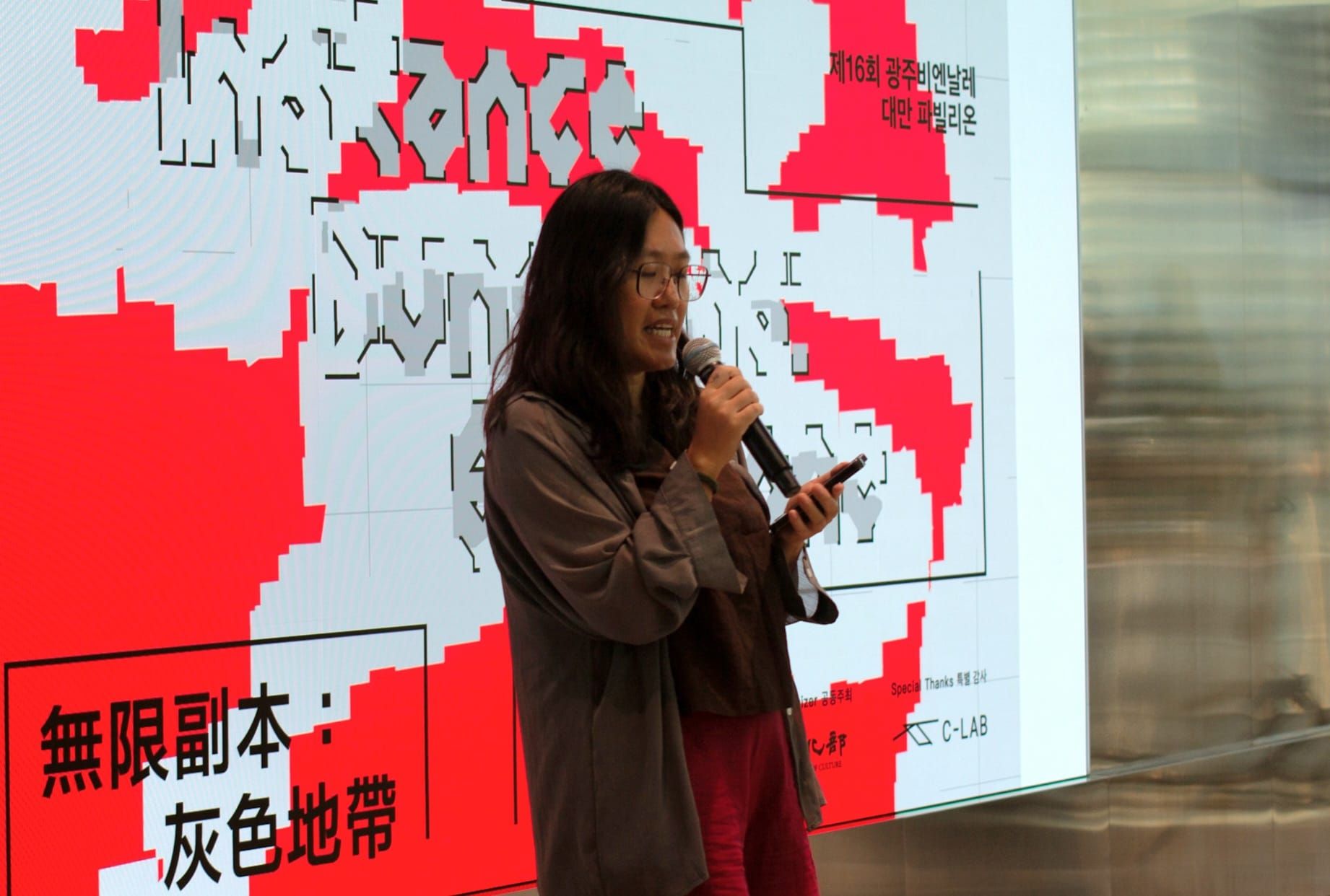 Ting-Ting Cheng holds a microphone and talks, looking down. Behind her is a screen with a graphic for the exhibition, reading, "Instance Dungeons: Gray Zone."