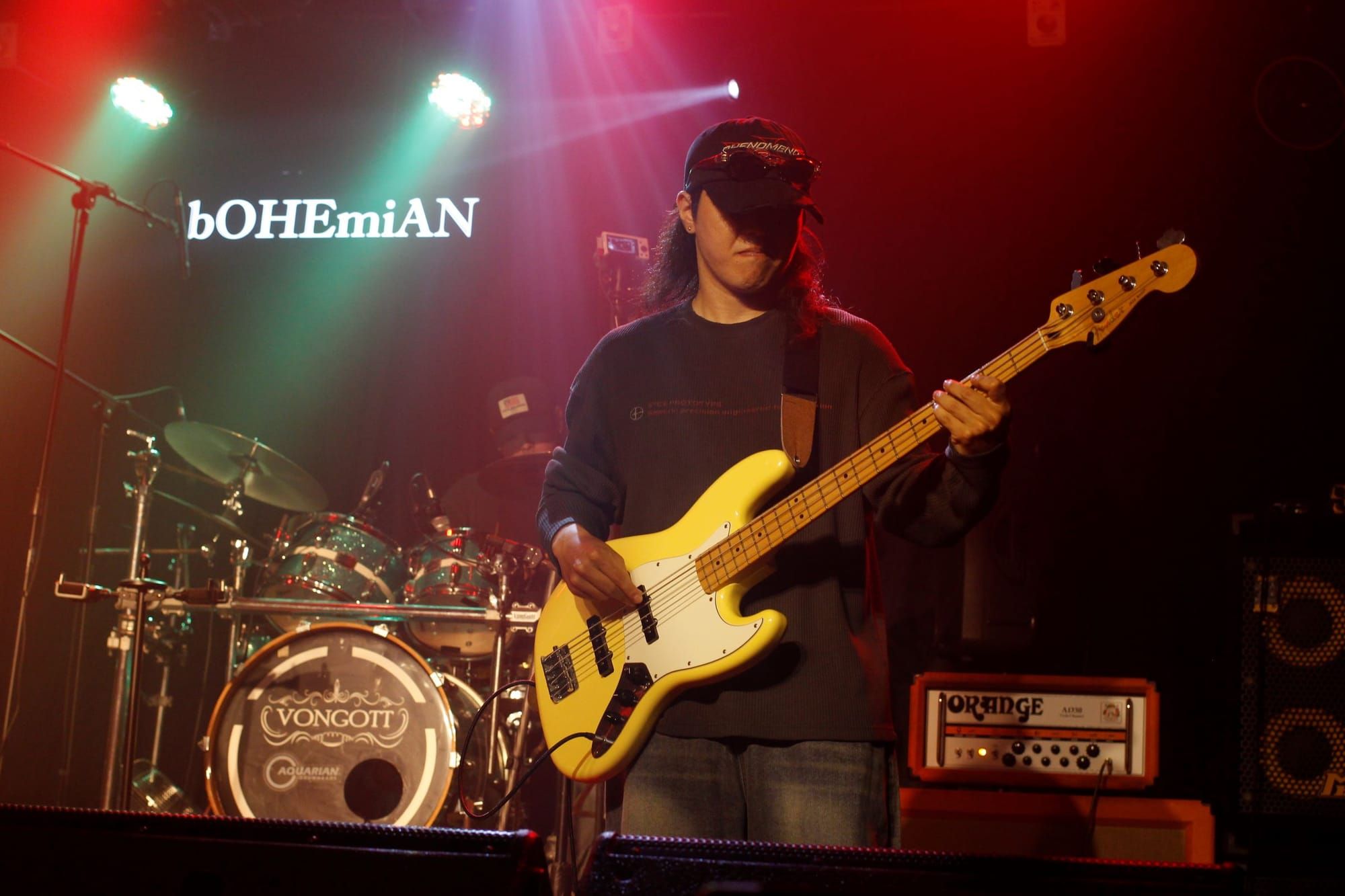 Shin Jaein frowns in a classic musical expression of pleasure while playing bass on stage. Behind him is the word bOHEmiAN and a drum set.