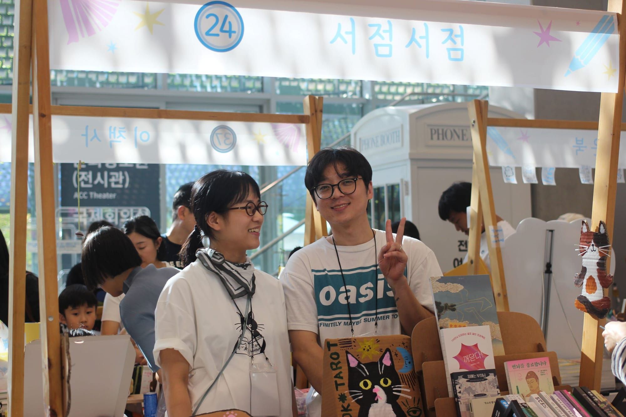 Yoon Nara smiles toward Seo Uihwa, who is facing the camera with a peace sign. They are wearing white shirts. A painting on cardboard shows a black and white cat with a star and moon.