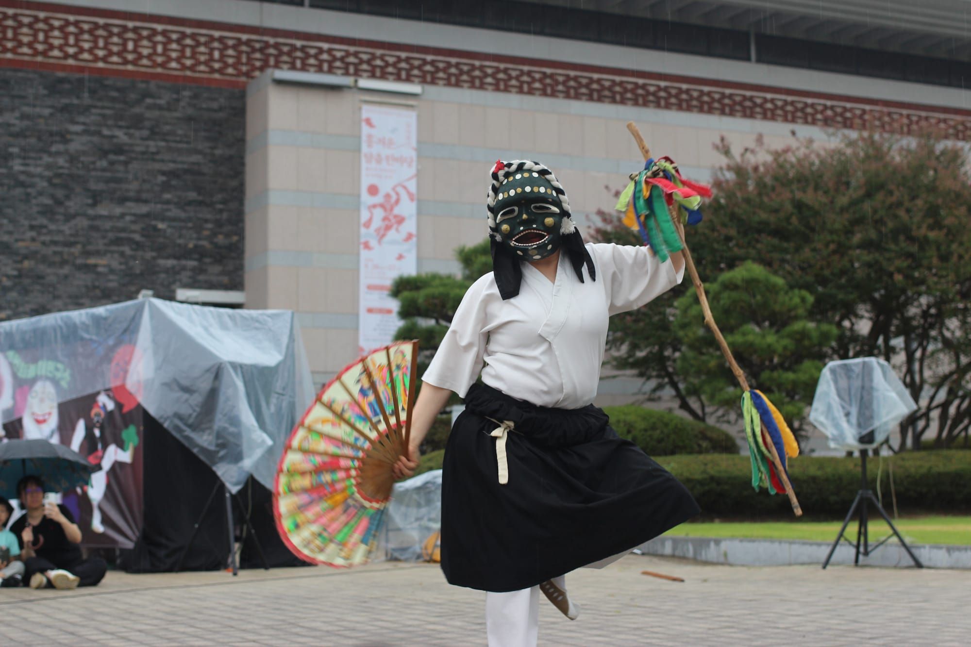 A dancer with a green folkloric mask, hand fan, and staff with ribbons dances in the same pose as a character in the banner behind her, on a museum pavilion.