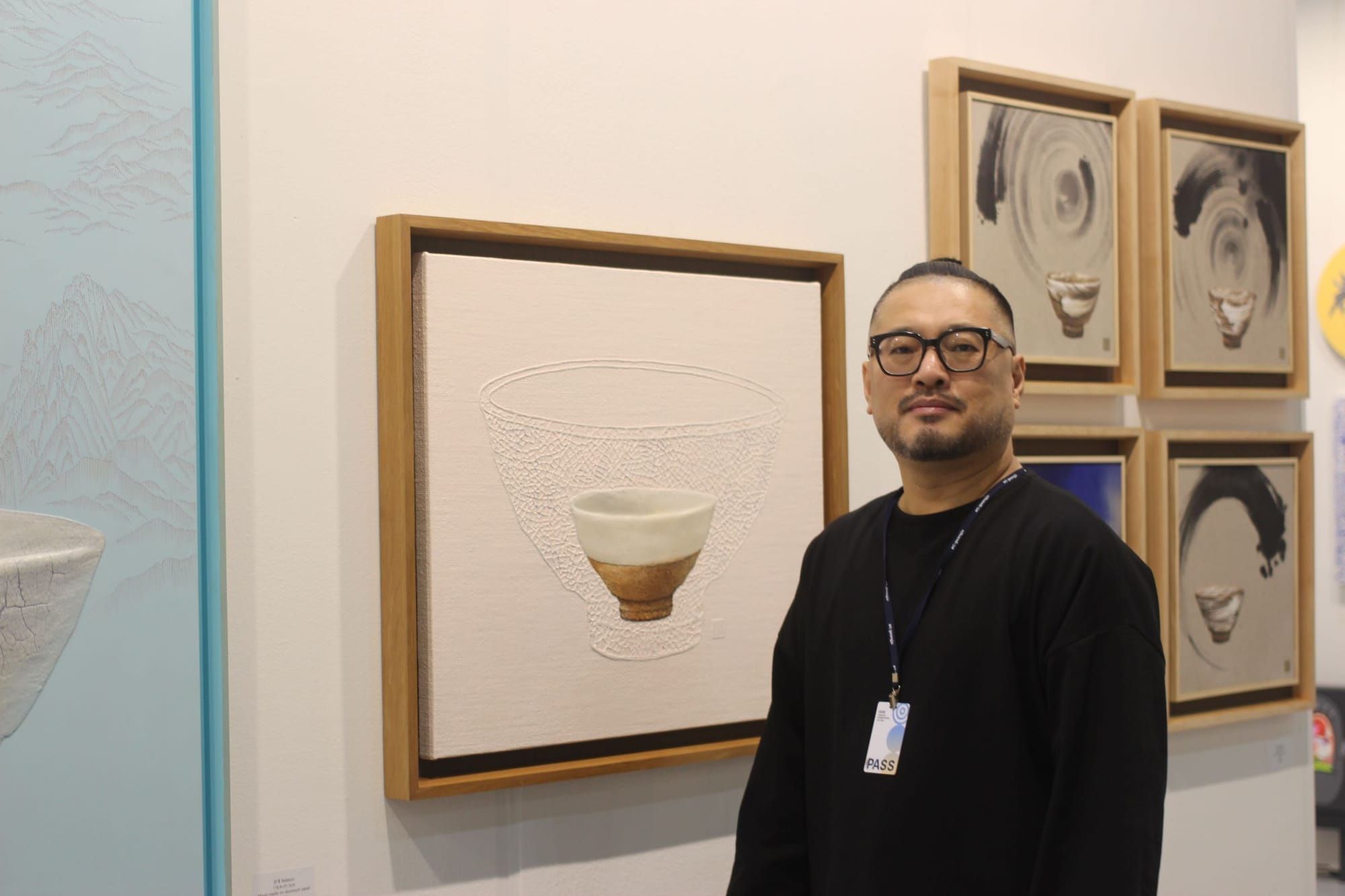 Seol Sang Ho looking toward the camera, in front of 5 square paintings of small ceramic bowls on large backgrounds.
