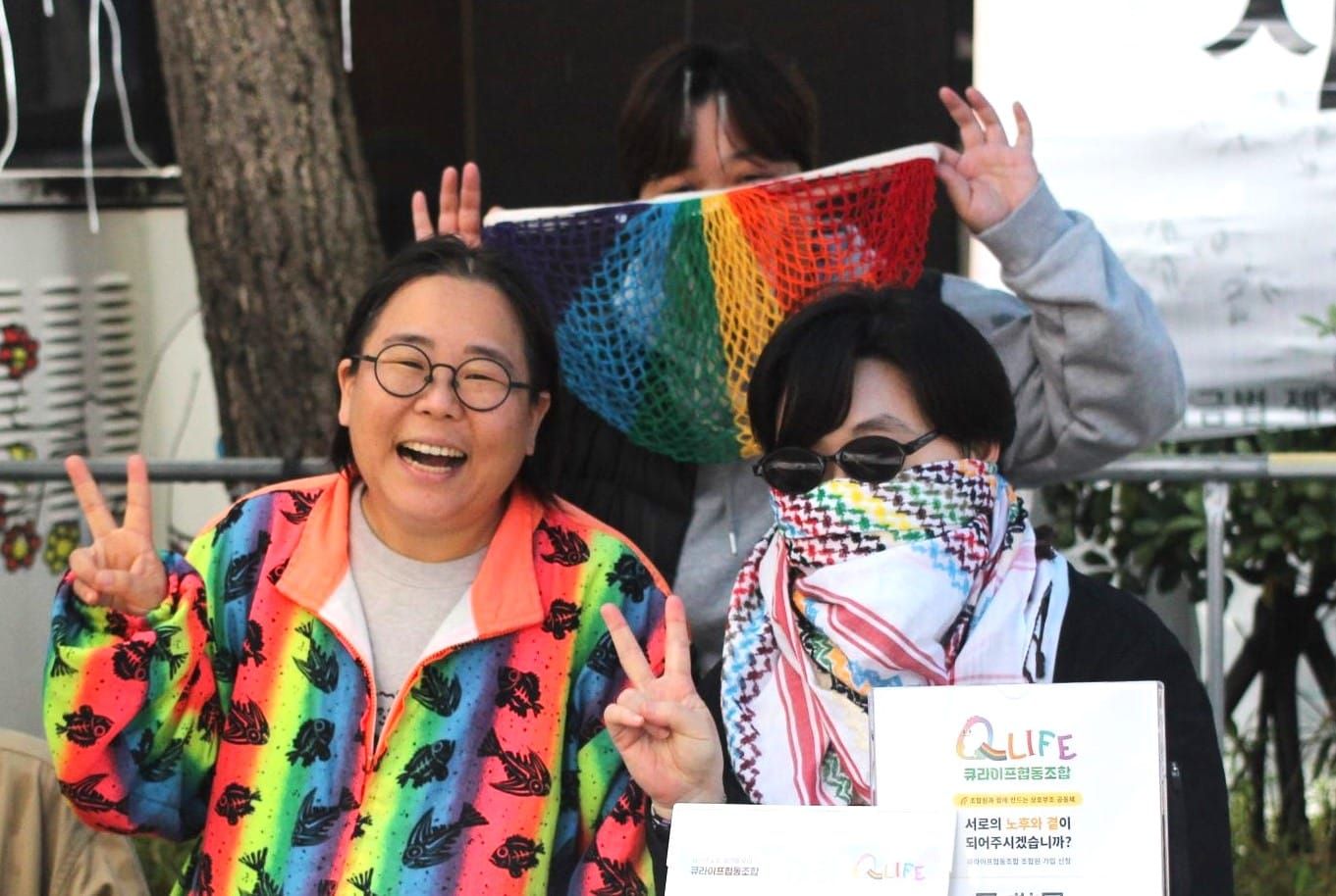 Three people close together, wearing rainbow colors and holding peace signs. The two younger ones have their faces partially covered.