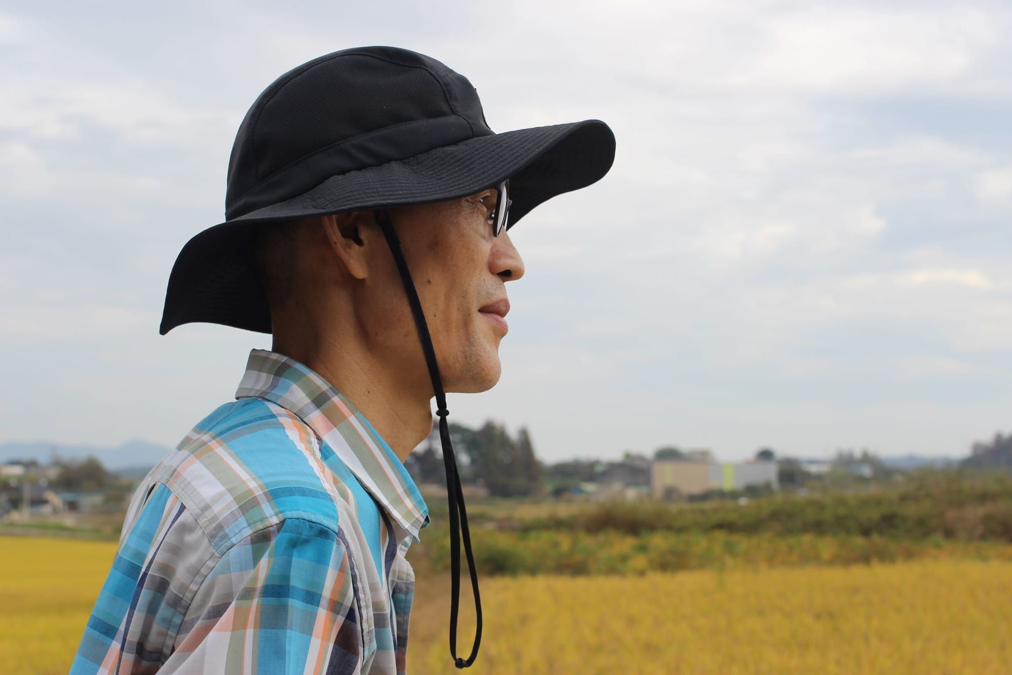 Yoon Hyung-Hyuk, wearing a hat with a brim, looks out over a yellow rice field.