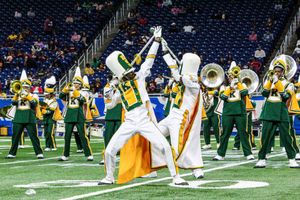 A marching band performs on a football field, with two members in white uniforms crossing their batons at the front while others in green and yellow play brass instruments. Photo credit: Akee