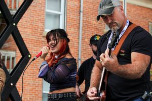 A woman sings into a red microphone while a man beside her plays an electric guitar outdoors. 