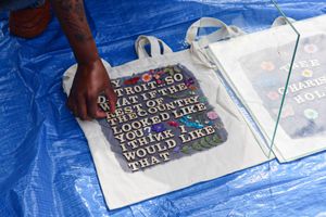 A hand arranges letters on a tote bag on a blue tarp that reads“Hey Detroit, what if the rest of the country looked like you? I think I would like that.”