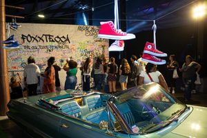 A crowd of people are facing a wall of graffiti behind a blue car, as shoes are tied to wires above.