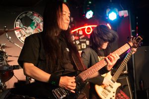 Lee Hee-Doo plays a guitar in a wide stance with a stoic expression. Behind him, bassist Han Seon-Jae flips his hair over his face while playing. Neon and stage lights illuminate them.