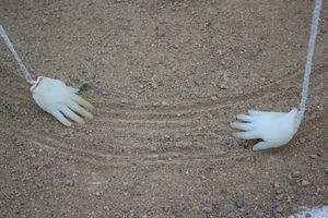 Two translucent hand sculptures suspended by white ropes touch the sandy ground, leaving grooves in their paths. Their fingers are pointed toward each other.