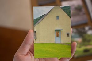 A hand holding a photograph of a simple triangular house in a bright green field. The house has small windows and a light blue door.