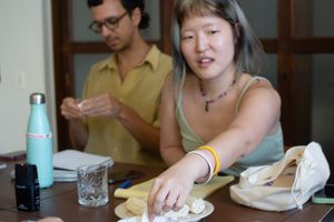 Romain, with a yellow collared shirt and glasses, reads a piece of paper in their hands. Jiann reaches forward, past a puffed rice snack, to select a paper.