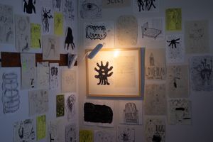 An interior corner filled with drawings on loose pieces of paper. A lamp illuminates a framed two-page spread featuring a monster with many eyeballs and a printed text passage.