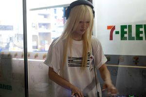 DJ Oto twists dials on a DJ deck with lit-up buttons. She has rings and bleached hair with straight-cut bangs. She is wearing big headphones.