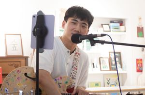 xx sits at a mic with a guitar covered in colorful drawings, with a guitar strap with prints of words like "Love," Kisses" and "XO." Behind are framed artworks.