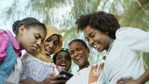 Five girls look at a phone screen in disgust and laughter. Behind is a tree; they are outside.