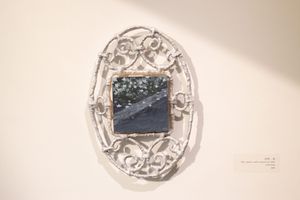 A small square painting framed by a textured white wire oval with curlicues. The painting depicts tiny white flowers and a cool gray solid surface, maybe asphalt.