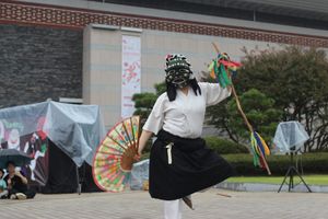 A dancer with a green folkloric mask, hand fan, and staff with ribbons dances in the same pose as a character in the banner behind her, on a museum pavilion.