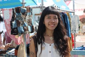 Kamonchanok Seansopha smiles, wearing a white outfit with light brown hand-dyed pattern, in front of other natural-dyed clothing.