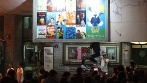 A large sign made up of 15 smaller paintings and one large painting of movie posters, hanging on the exterior of a building. From behind, a man is climbing down using a ladder.