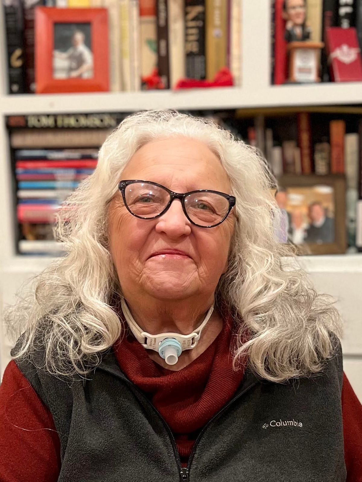 An woman with shoulder-length wavy gray hair smiles at the camera. She is wearing glasses, a rust coloured turtleneck and a dark gray fleece vest.  She sits in front of a bookcase. She has 