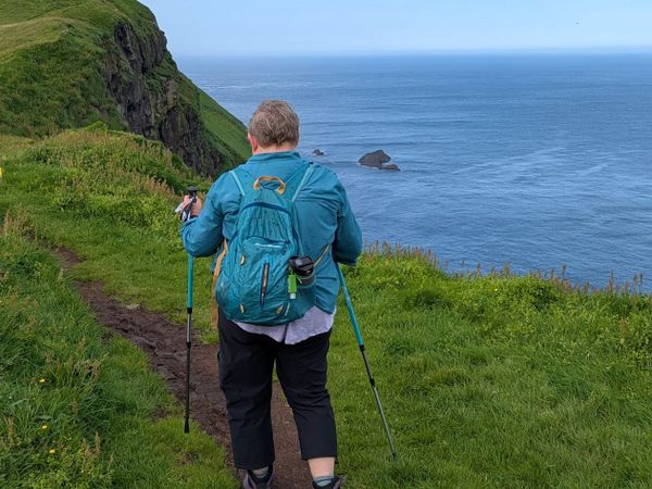 A woman pictured from behind, hiking along a pathway that overlooks the ocean.  She is using turquoise hiking poles and is wearing a teal backpack.  
