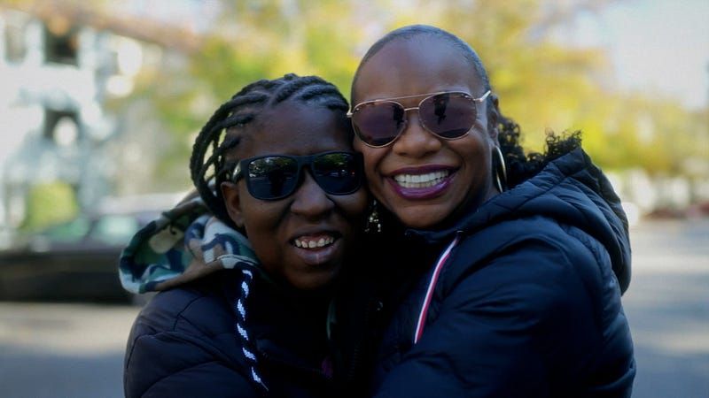 Two women of color with sunglasses and winter coats hugging outside with trees behind them.