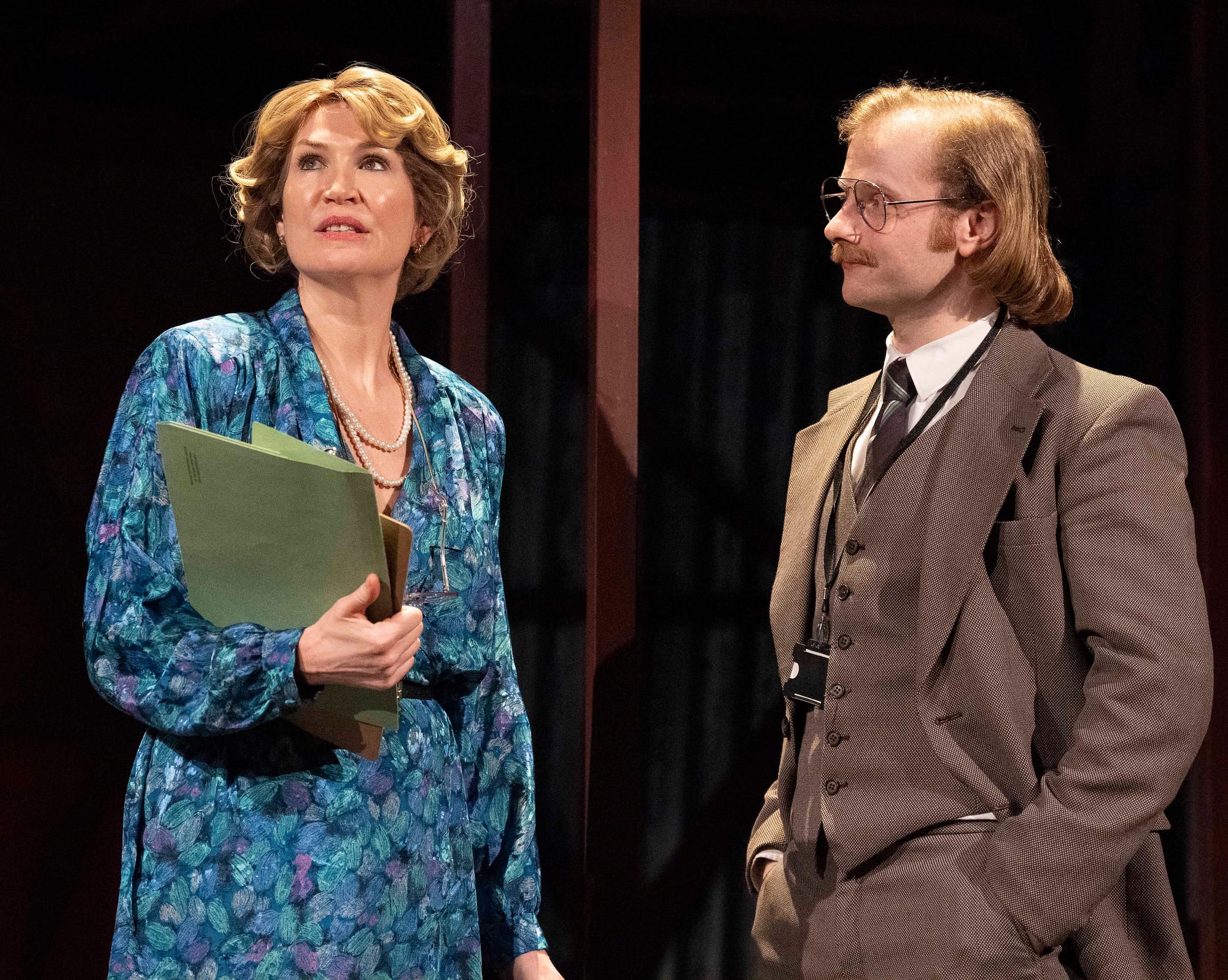 A perfectly coiffed woman in an absolutely divine blue floral ensemble clutches some papers while engaging in what appears to be very important theatrical business with a bespectacled gentleman. Their 1980's  period costumes and intense expressions suggest discussions importance.