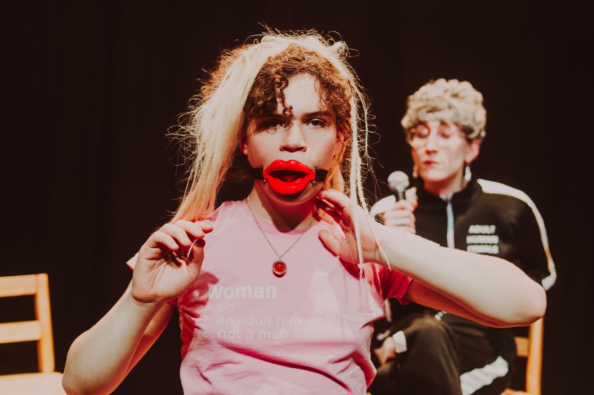 A performer with curly hair with rough blonde extensions wearing a pink t-shirt that reads 'Woman' looks out at us and wears a red mouth gag lip piece. Another performer, holding a microphone, with glasses and a black sports suit,  is visible in the background. The lighting creates a warm, intimate atmosphere typical of experimental theater.