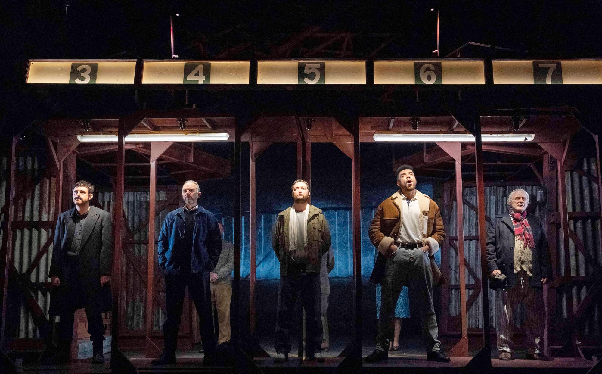 Five devastatingly handsome men stand dramatically lit in a post industrial setting, numbered compartments glowing above them like some sort of testosterone lottery system. Their brooding poses suggest they're waiting. .