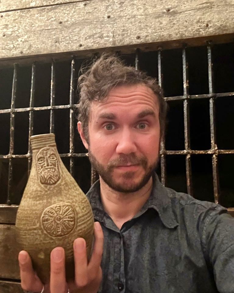 A man with tousled curls poses dramatically with an ornate ceramic vessel, against a backdrop of weathered stone and wrought iron 