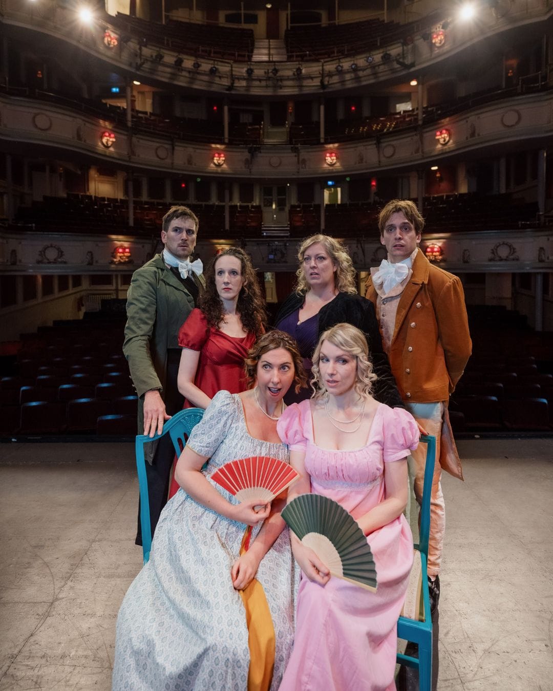 Six actors in Regency costumes pose theatrically in an ornate theater - two women with fans sit front, four others stand behind in period dress. , it's giving Jane Austen meets drama club realness! Fan-fluttering ladies serving ethereal vibes while the gents bookend this Regency fantasy! 