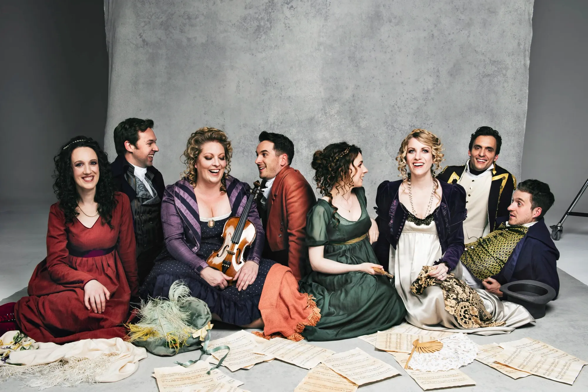 Eight actors in Regency costumes sit on floor surrounded by scattered sheet music and props, including violin and feathered hat. it's giving bohemian artist salon realness! Our Regency squad is serving intimate musical soirée with violin, scattered manuscripts, and pure theatrical chaos!