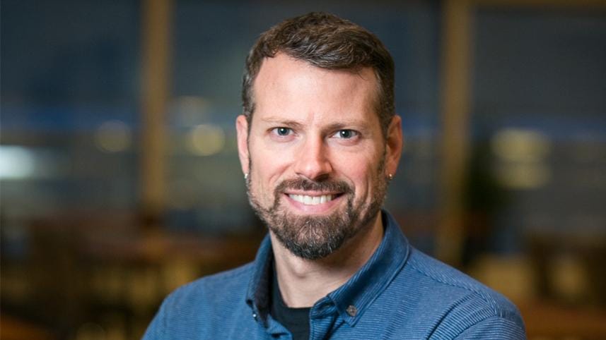 Professional headshot of a smiling man with brown hair and a well-groomed beard wearing a blue collared shirt. The background shows a blurred office or business environment with warm lighting